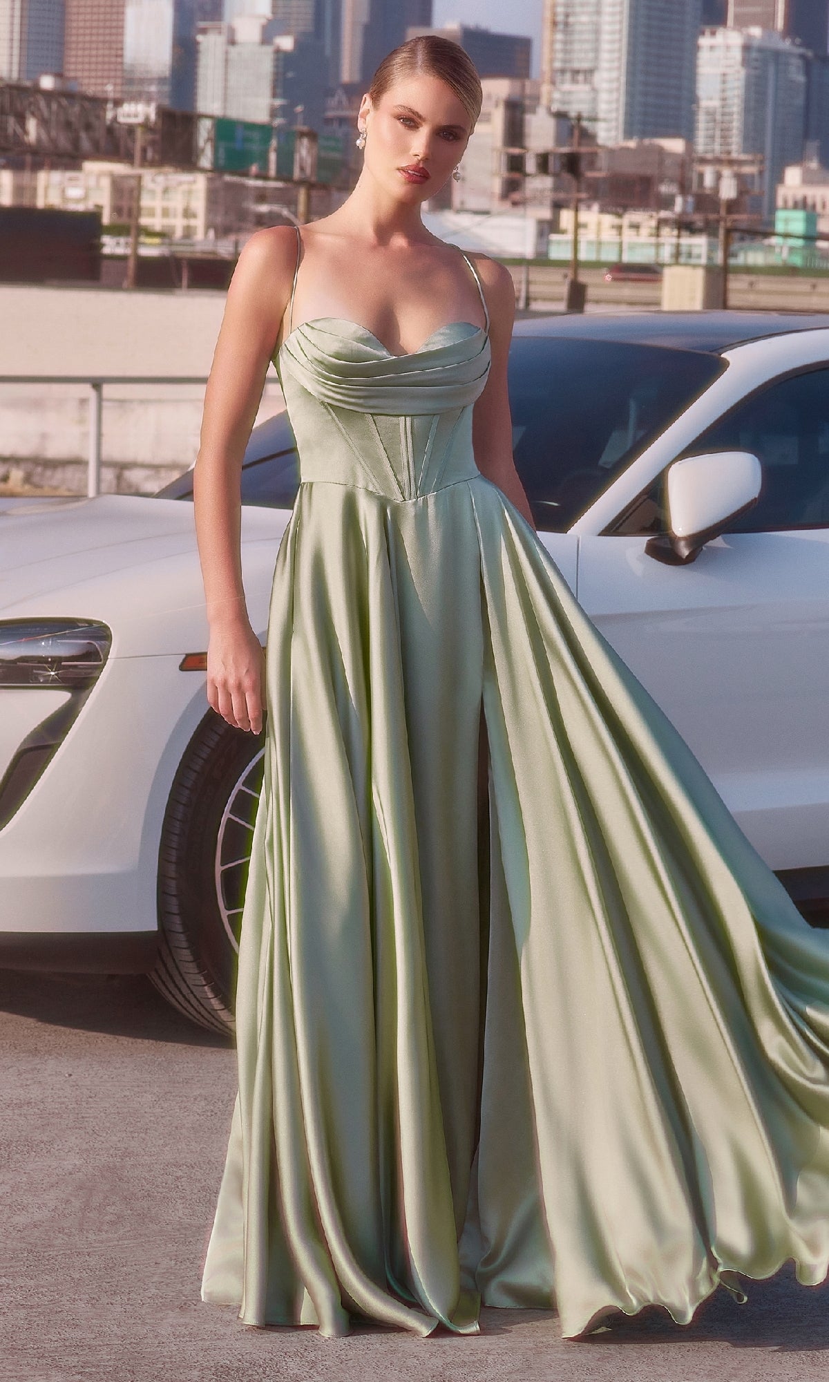 A woman wearing the Ladivine 7481 long satin A-line prom dress in pale green stands before a white car with a cityscape behind her.